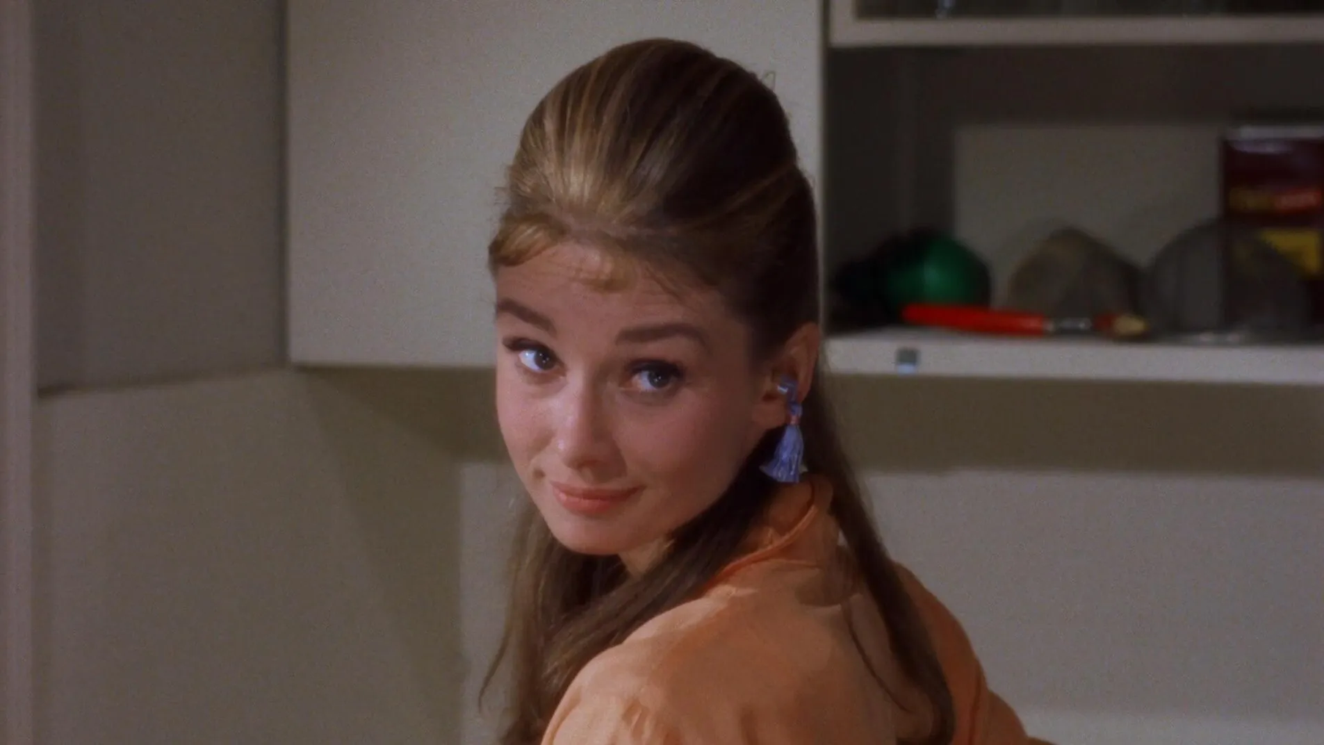 Audrey Hepburn, as Holly Golightly, looks back over her shoulder towards us. She is wearing an orange dressing gown and purple earplugs with fringe dangling from them. Audrey Hepburn, as Holly Golightly, looks back over her shoulder towards us. She is wearing an orange dressing gown and purple earplugs with fringe dangling from them.